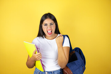 Young beautiful student woman wearing backpack holding notebook over isolated yellow background very happy and excited making winner gesture with raised arms, smiling and screaming for success.