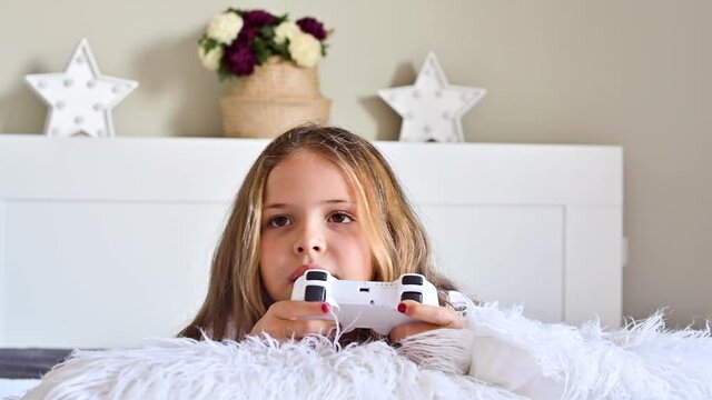 A little girl is playing a computer console on a sofa in a bright room. Concurrent games for children. Joystick for control in hands. Emotions and fun of the child. Copy space.High quality 4k footage.