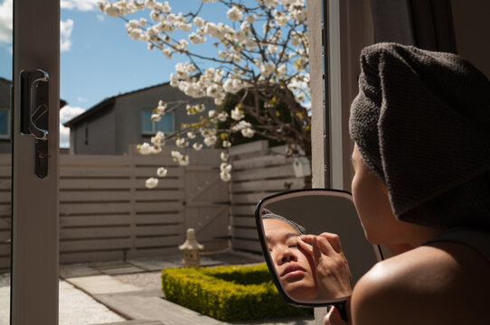 An Asian girl's face with a towel over her head is reflected in a hand mirror as she applies make up in her eye in front of a zen looking garden with a cherry blossom tree and a hedge in Edinburgh, UK