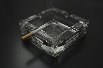 glass ashtray with burning cigarette and smoke on a black ground