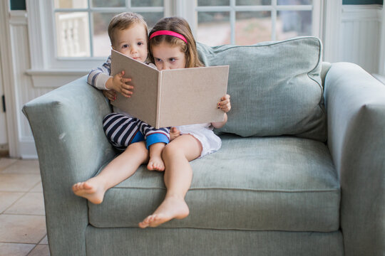 Young girl and boy toddler sitting on an oversized chair reading a book