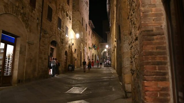 San Gimignano,Tuscany,Italy. August 2020. The nocturnal charm of the medieval-looking alleys: stone-paved street and brick houses,the yellowish light weakly illuminates the street,people walk along it