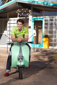 Portrait of man sitting on vespa outside a mid century style home. 