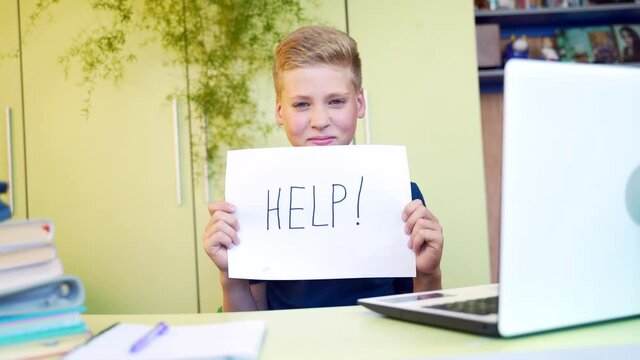 portrait of a tired young blond boy teaches lessons with a notebook and laptop at table in the home. Teen schoolboy shows a picture with the inscription help while studying. Close-up face exhausted