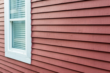 A small closed glass double hung window with white trim. There's a white blind hanging from the inside of the window. The exterior wall of the building is made of bright red narrow wooden clapboard.