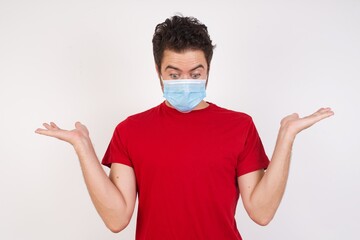 Hesitant  Young caucasian man with short hair wearing medical mask standing over isolated white background shrugs shoulders, looks uncertain and confused. Have no answer