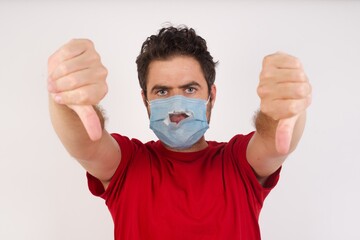 Young caucasian man with short hair wearing broken medical mask standing over isolated white background being upset showing thumb down with two hands. Dislike concept.