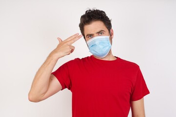 Unhappy  Young caucasian man with short hair wearing medical mask standing over isolated white background  makes suicide gesture and imitates gun with hand, curves lips, keeps two fingers on temple