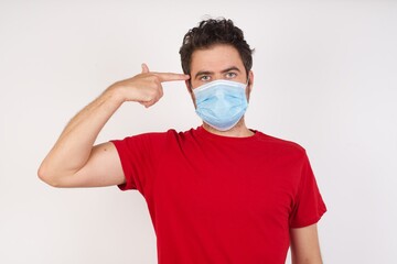 Unhappy  Young caucasian man with short hair wearing medical mask  makes suicide gesture and imitates gun with hand, curves lips keeps two fingers on temple, shoots, being tired of everything,