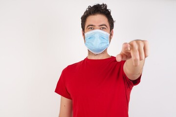 Young caucasian man with short hair wearing medical mask standing over isolated white background pointing at camera with a satisfied, confident, friendly smile, choosing you