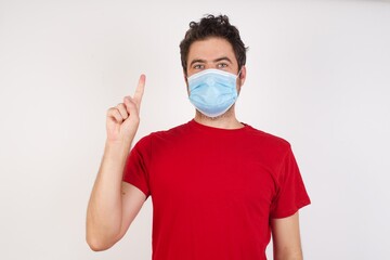 Young caucasian man with short hair wearing medical mask standing over isolated white background showing and pointing up with finger number one while smiling confident and happy.