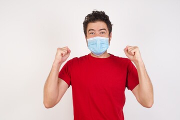 Young caucasian man with short hair wearing medical mask standing over isolated white background celebrating surprised and amazed for success with arms raised and open eyes. Winner concept.