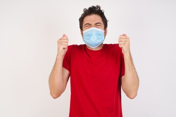 Young caucasian man with short hair wearing medical mask standing over isolated white background being excited for success with raised arms and closed eyes celebrating victory. Winner concept.
