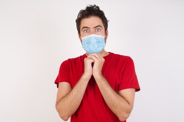 Young caucasian man with short hair wearing medical mask standing over isolated white background praying for luck has hands crossed near face, amazed and opening mouth looking front.