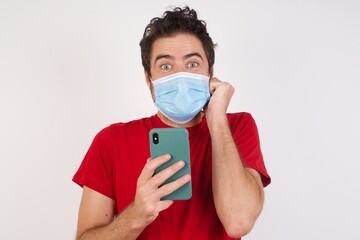 Joyous Young caucasian man  wearing medical mask poses with mobile phone device, types text message on modern smartphone, watches funny video during free time, enjoys good internet connection,