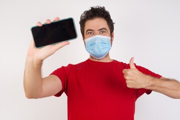 Young caucasian man with short hair wearing medical mask standing over isolated white background holding in hands cell showing giving black screen thumb up
