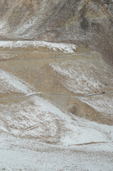 Patches of snow cover a rocky mountain slope in Ladakh, India. A road winds across it. A truck is just visible, dwarfed by the size of the mountain.