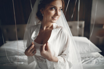 Beautiful bride sitting on a bed. The girl in a white pajamas and veil