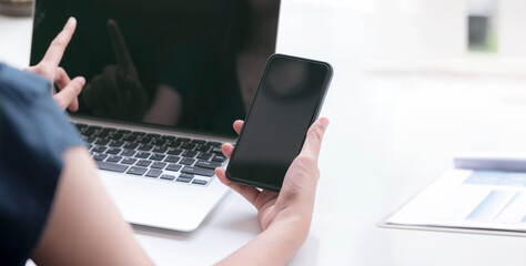 Cropped shote of female hand holding blank screen smartphone while sitting at the table and working with laptop.
