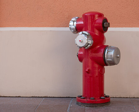 A fire hydrant for extinguishing water stands in front of a house wall. The object is painted red and has silver screw caps. The base is screwed into the floor.