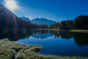 Sun on a crystal blue sky. Early morning in the Alps. House in the mountains by a mountain lake. Reflections of trees, mountains, sky in a mountain lake.