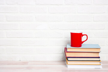 Fototapeta na wymiar Stack of books and red mug against a white brick wall.