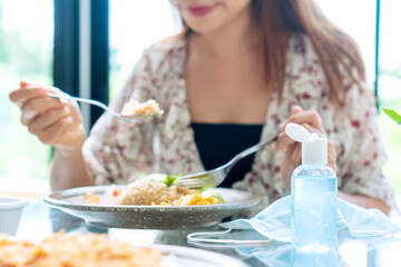 Beautiful young Asian woman have lunch and sanitizer gel, surgical face mask on table in restaurant. New normal lifestyle, preventive measures during the period of pandemic concept. Closeup