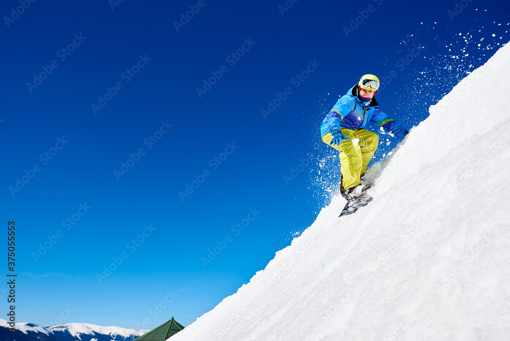 Wall mural male snowboarder riding snowboard fast down steep snowy mountain slope, jumping in air on copy space