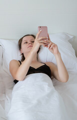 Woman in bed with cellphone watching latest new. Young adult girl lying in white sheets bed in the...