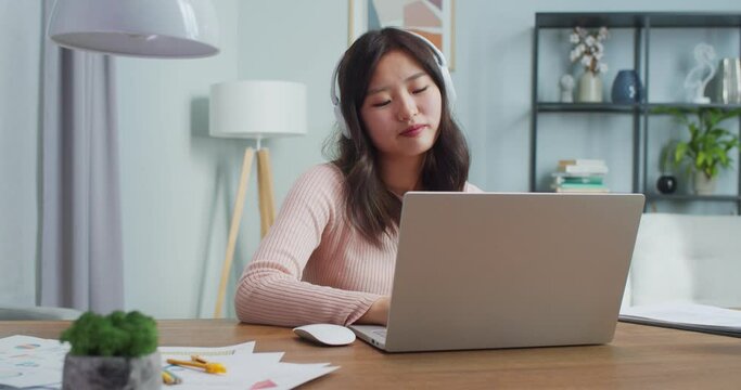 Pretty Asian woman sitting at table and working, studying in home office. Relaxed young girl in headphones using laptop and shaking head while listening to music. Internet, distance learning.