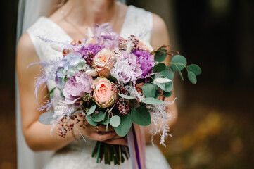 Young beautiful girl in elegant dress is standing and holding hand bouquet of pastel pink flowers and greens with ribbon at nature. The bride holds a wedding bouquet outdoors.