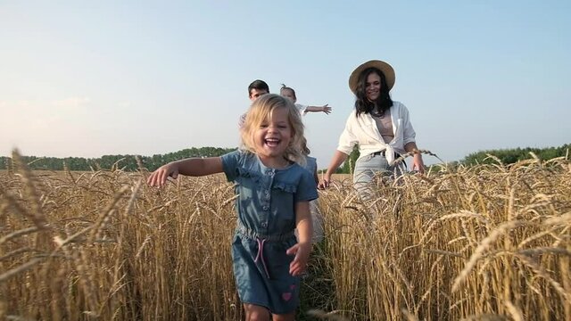 Caucasian large family with mother and three daughters walking in field at sunset. Happy parents play with their children outside the house. little girl in field runs to the camera and laughs merrily.
