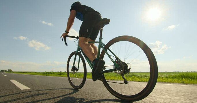 A sportsman on a bicycle is standing on the pedals and developing great speed. Training on a bicycle. Shooting from behind. Sun in the background. 4K