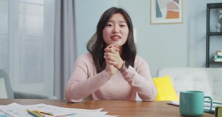 Pretty young Asian woman sitting at table in modern minimalist room at home. Smiling female vlogger is talking to camera, making live conference, recording blog. Social media, vlogging.