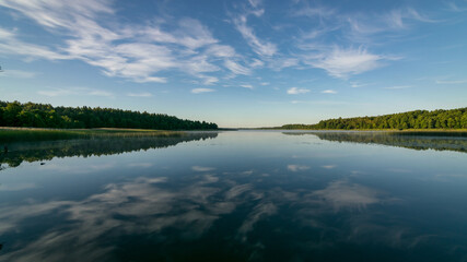 cloud reflections in clear and calm lake water, forest in the background, summer morning