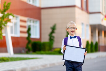 a happy schoolboy boy with blond glasses and a backpack stands at the school and holds a sign with a white sheet. Day of knowledge. Space for text