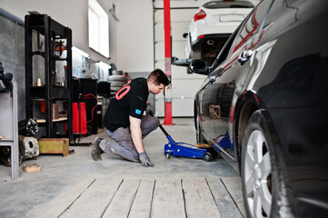 Car repair and maintenance theme. Mechanic in uniform working in auto service, lifted with hydraulic floor jack for repairing.