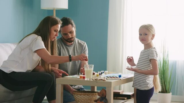 Young parents, mom and dad, are painting with their small daughter, using watercolors, having greate time together, enjoying common hobby, girl makes funny face and looks at camera, Slow motion.