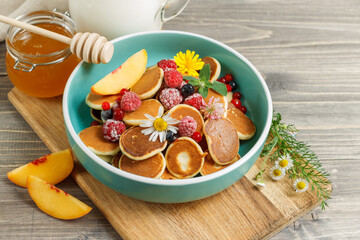 mini pancakes with fruit and berries in a blue bowl, decorated with fresh flowers. Close-up on a wooden surface, horizontally