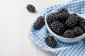 Top view of white bowl with blackberries on blue checkered cloth, on white wooden table, horizontal, with copy space