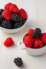 Top view of two white bowls with blackberries and raspberries on white wooden table, vertical, with copy space