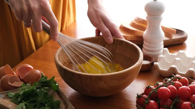 Mixing eggs with a wire whisk in wooden bowl. Woman prepares scrambled eggs or omelette for breakfast