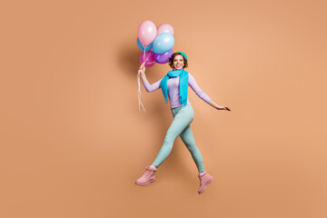 Full body profile photo of pretty lady jump high carry many air balloons walk surprise party wear violet jumper green pants boots blue beret scarf isolated beige color background