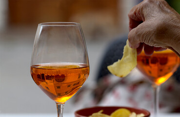 Elderly wrinkled hand holding potato chips and two glasses of delicious Aperol Spritz cocktail on blurred background from Venice