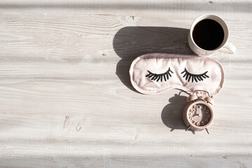 sleep mask, coffee cup and alarm clock on a wooden white table in the morning sunlight. The concept of starting a new day, good morning or ending the day, late evening. Business planning