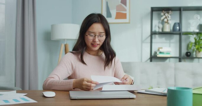 Pretty Asian woman has recieved response from university, college. Excited young girl in glasses is sitting at table and opening letter. Emotional student being sad of refusal and taling off glasses.