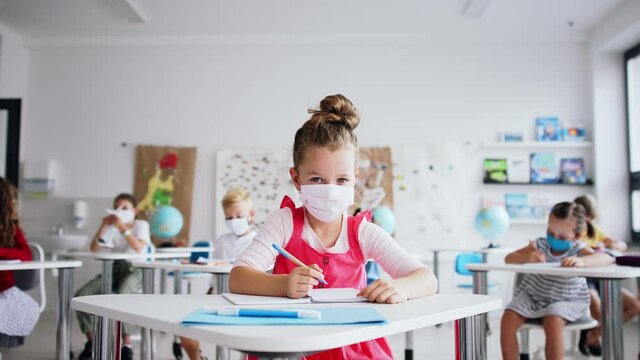 Small girl with face mask sitting on lesson indoors in classroom, looking at camera.