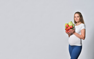 Pregnant blonde woman holding fresh vegetables in bowl on gray background