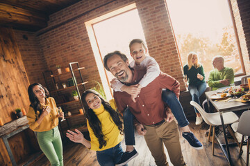 Photo of full family gathering six people two small kids play dad piggyback daughter wave hand mom laugh grandparents sit sofa dinner table generation in home evening living room indoors