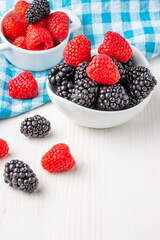 Top view of two white bowls with blackberries and raspberries on blue checkered cloth, on white wooden table, vertically, with